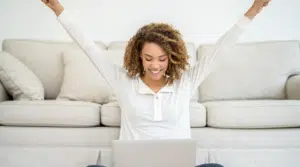 Une femme souriante devant son ordinateur, célébrant la réussite de son projet pour générer un complément de revenus depuis chez elle.